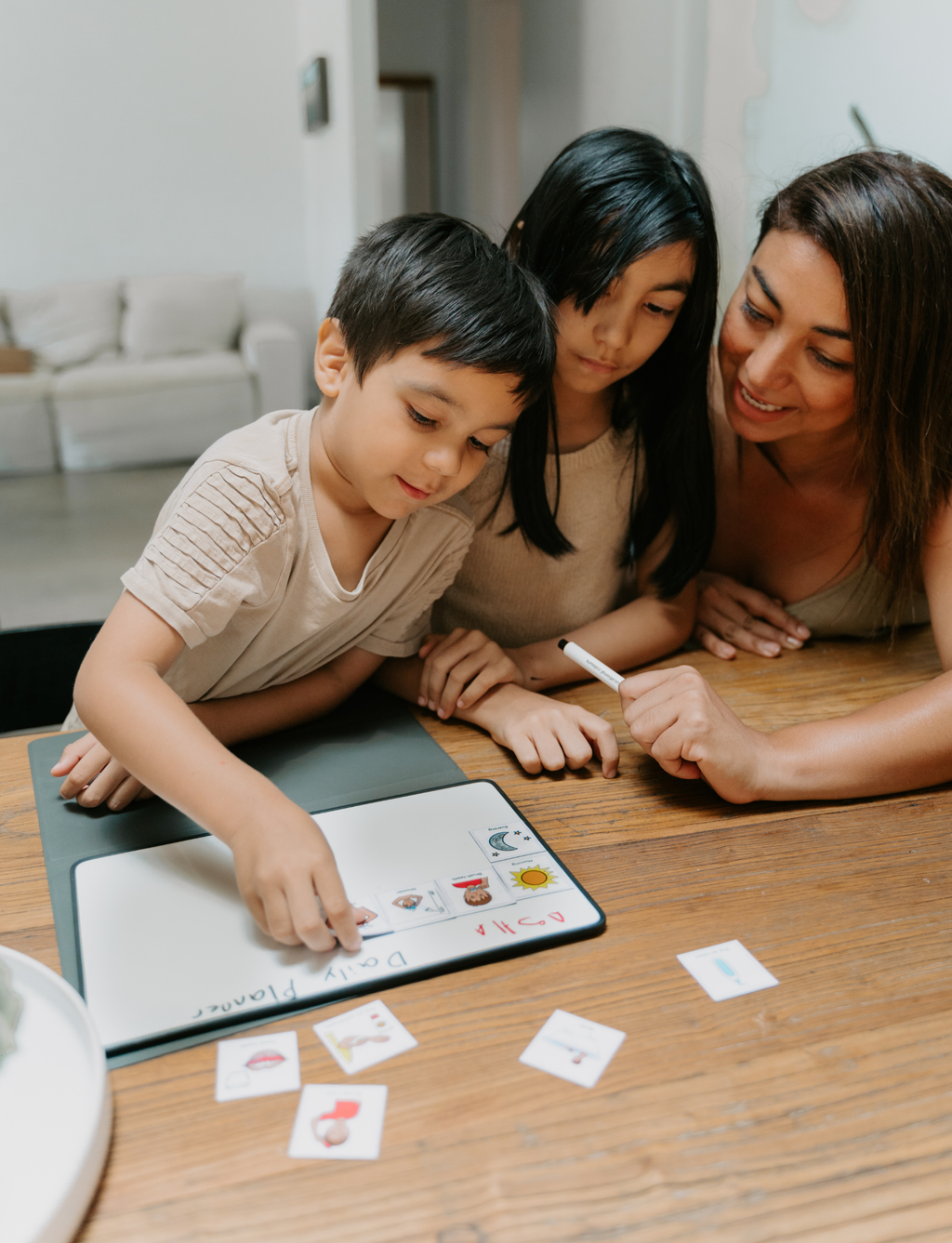 A mother and two children are gathered around a tablet on a wooden table, interacting with colorful educational cards and a whiteboard marker in a bright, modern home setting.