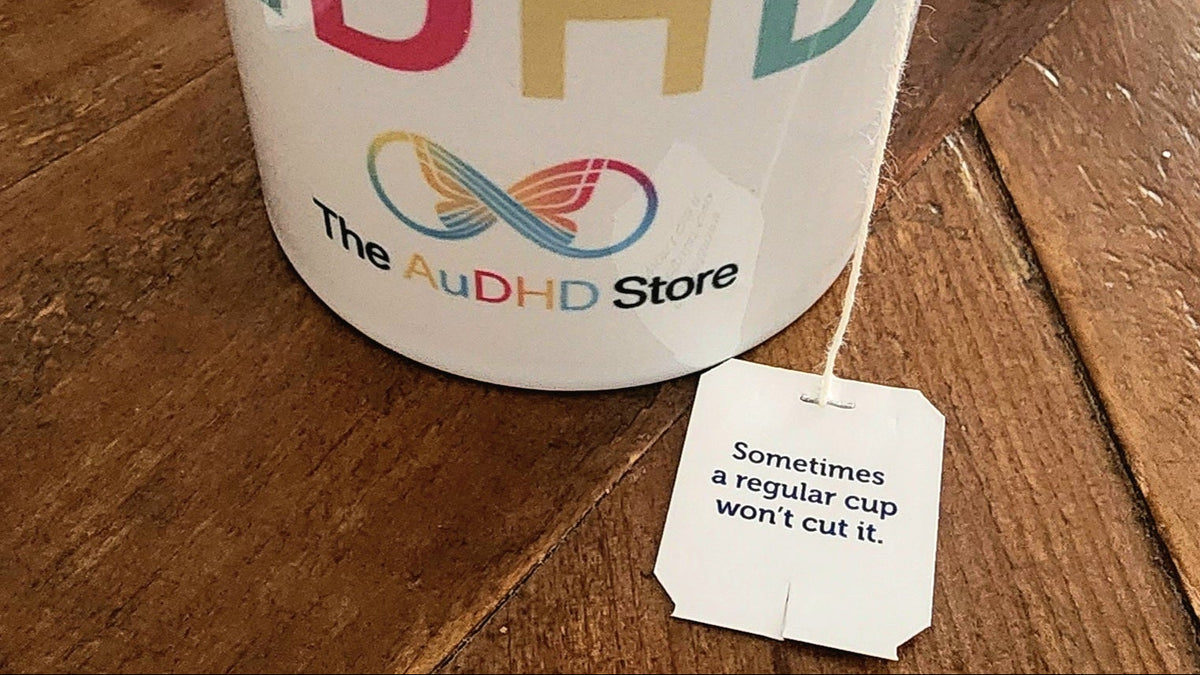 A white ceramic mug filled with tea sits on a wooden table. The mug is printed with the text 'AuDHD' in colorful letters and 'The AuDHD Store' logo below it, with a tea bag tag hanging from the side.