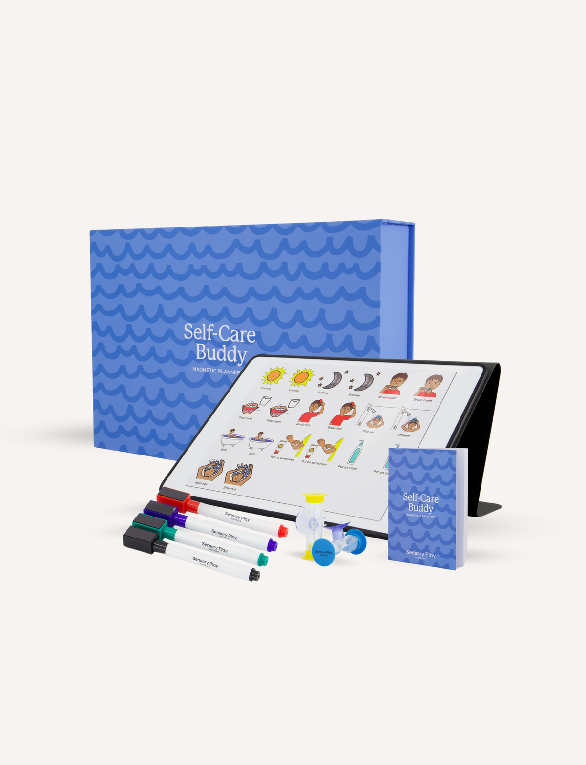 Self-Care Buddy therapy kit displayed on a clean off-white background, featuring a tablet with illustrated self-care options, a blue patterned storage box, a small blue booklet, and several colored markers.