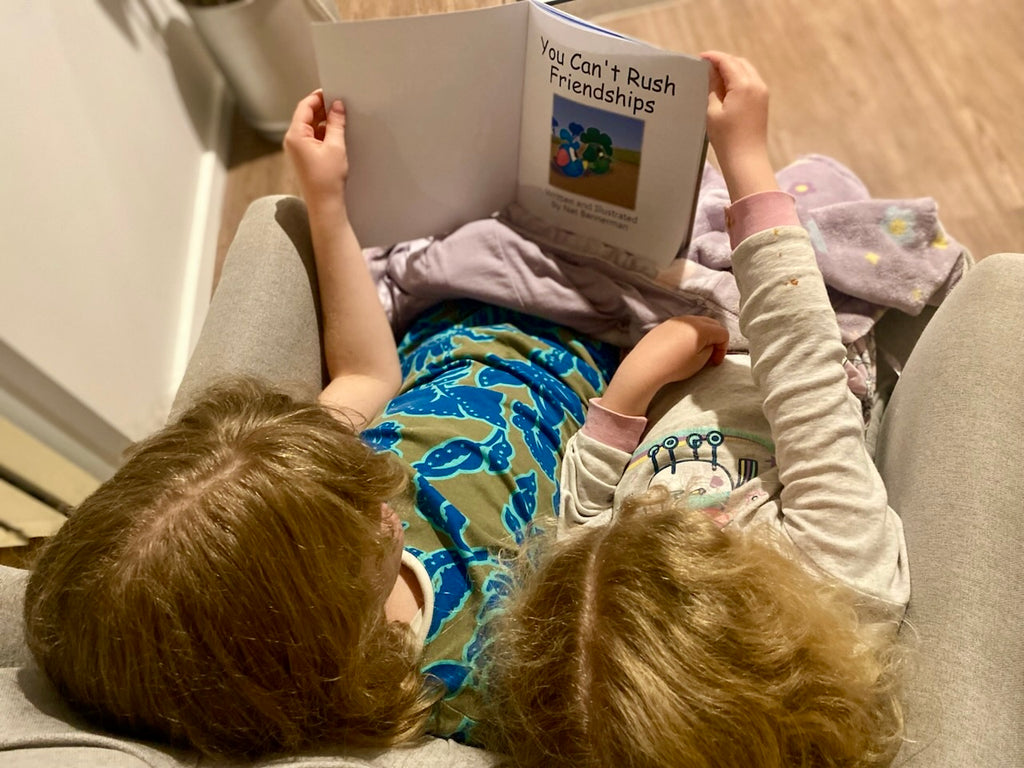 Two children lying on a gray couch reading a book titled 'You Can't Rush Friendships'.
