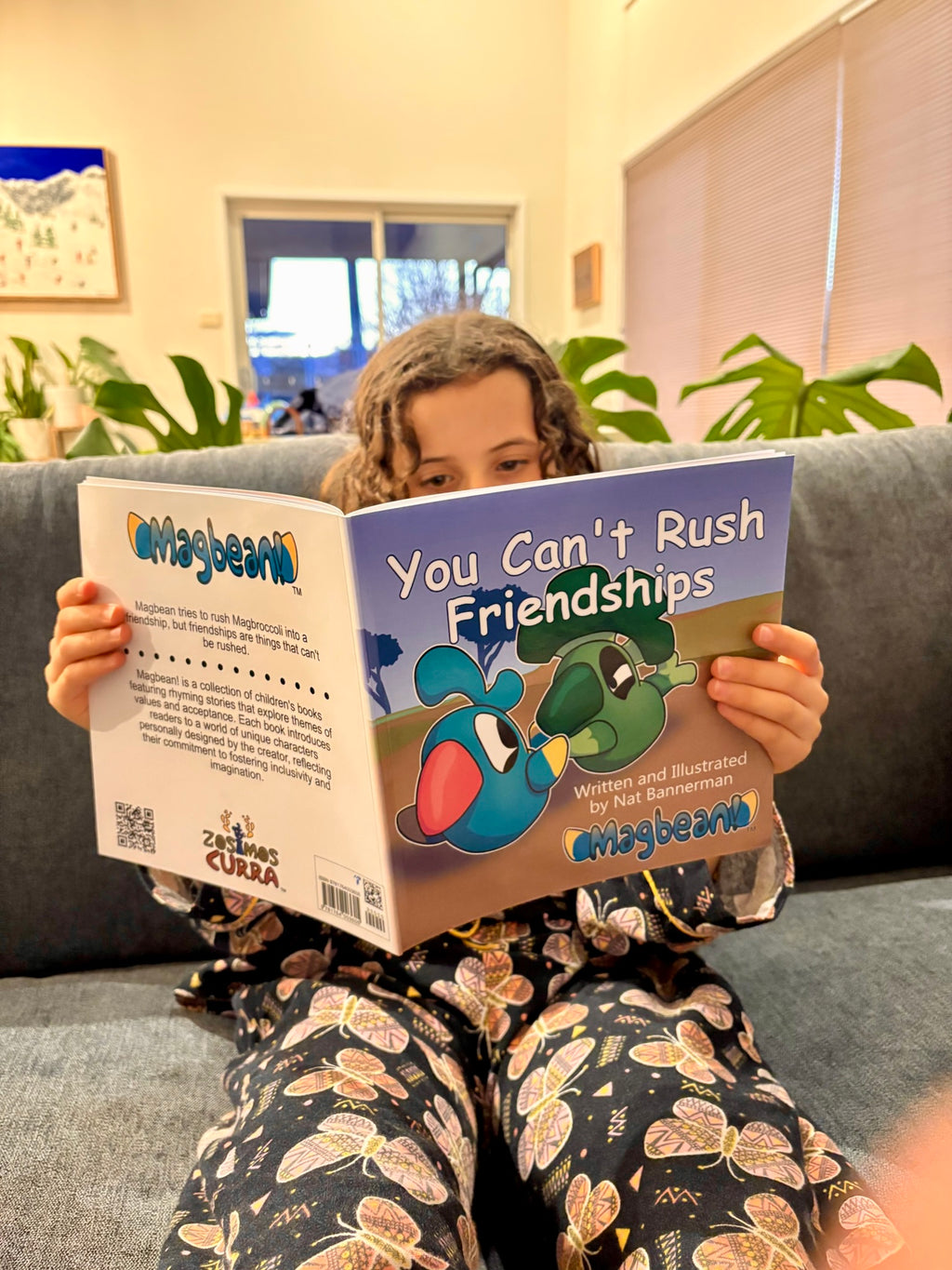 A child sitting on a gray couch holding a book titled 'You Can't Rush Friendships' by Ariagbean, with colorful cartoon illustrations on the cover.