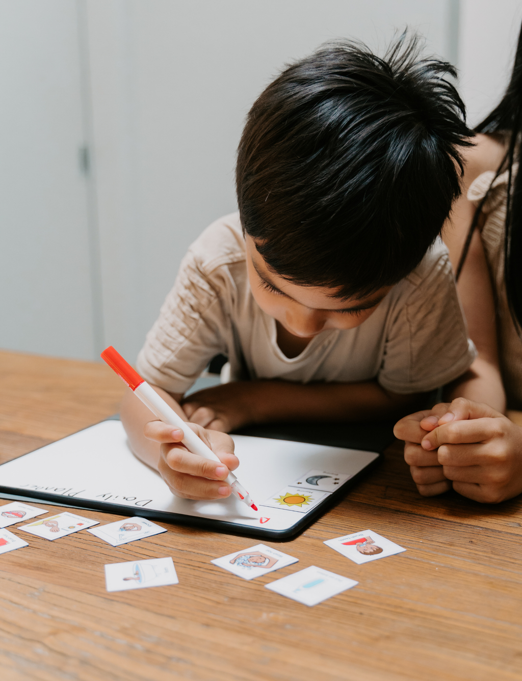 A young boy with dark hair leans over a tablet on a wooden table, using a red marker to draw. Small image cards are scattered around the table, and part of an adult is visible behind him.