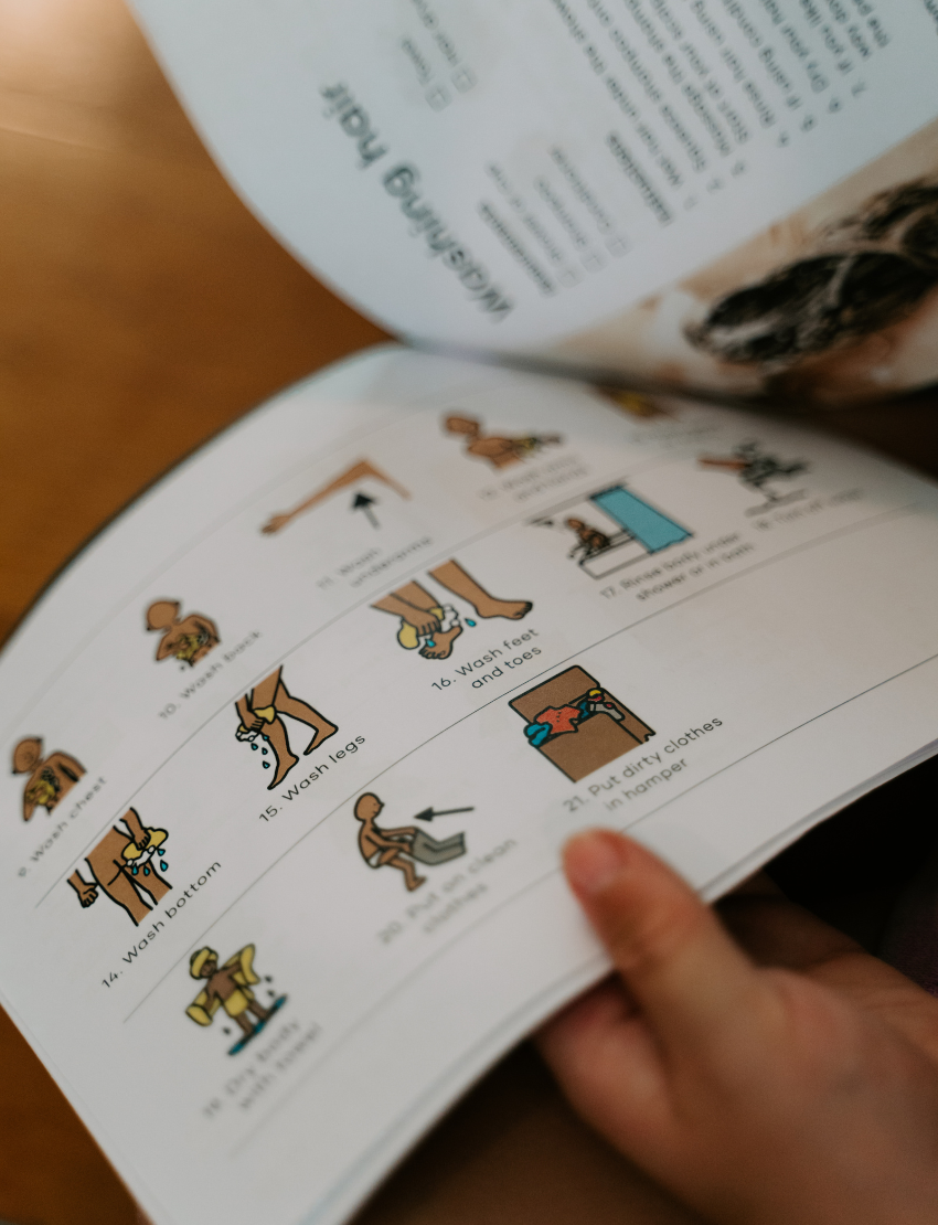A hand holding an open booklet or instruction manual on a wooden table, showing illustrated steps for grooming or care.