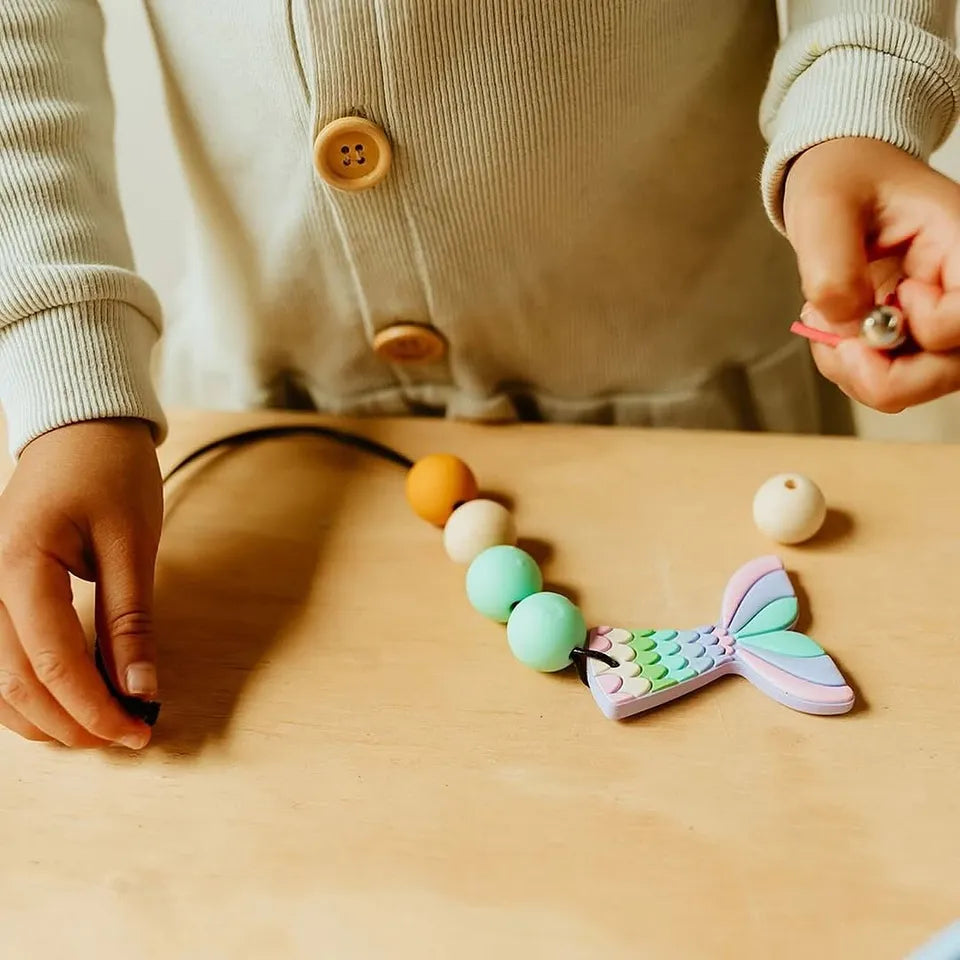 Colorful mermaid tail teething toy on a wooden surface with a person's hands nearby.